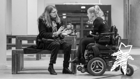 Two women talking. One woman is sitting on a bench. The other woman is in a wheelchair.