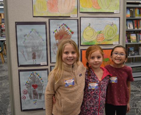 three girls stand in front of an art board displaying colorful artwork