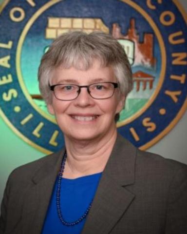 Official Portrait of Maggie Trevor wearing a gray suit with a royal blue blouse in from of a county seal. She wears her gray hair short and wears glasses.