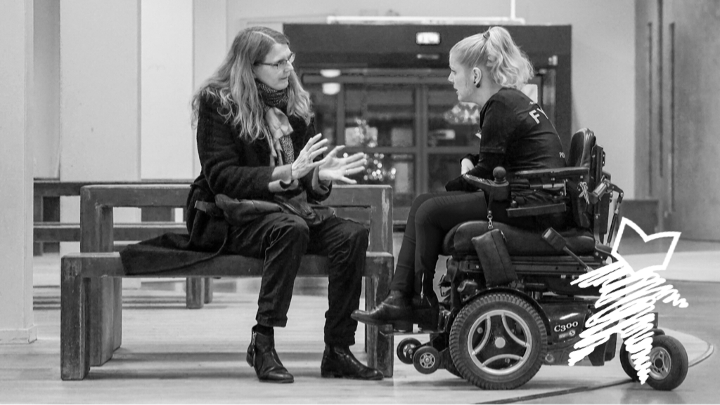 Two women talking. One woman is sitting on a bench. The other woman is in a wheelchair.